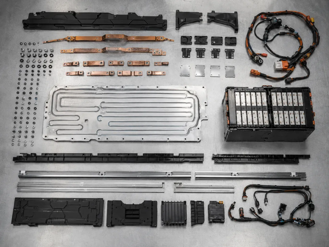 The recovered components of one battery pack laid out in ordered rows on a steel bench: busbars, cooling plate, fasteners, wiring harness and an intact module