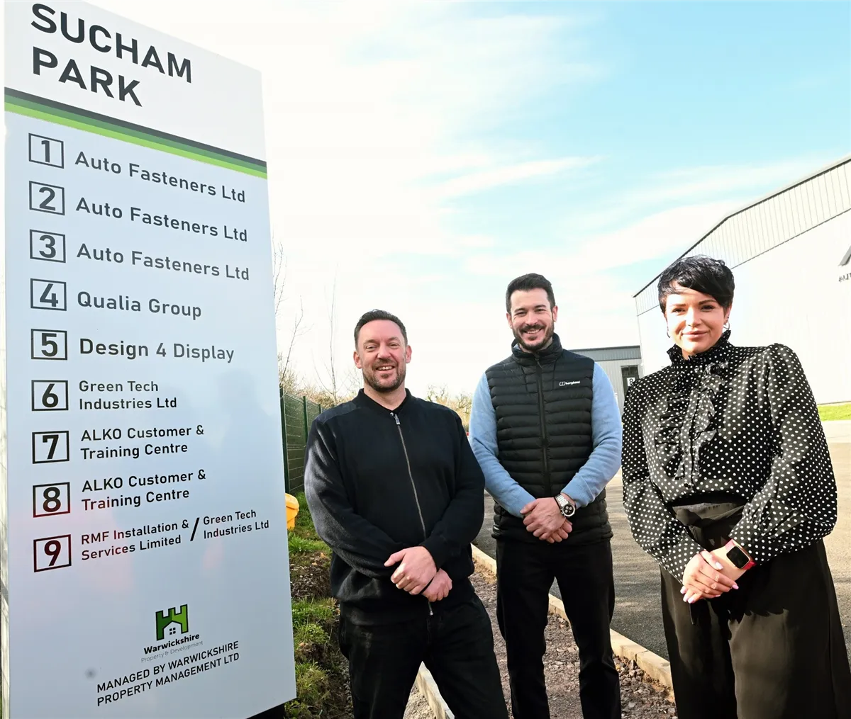 Three people beside the Sucham Park entrance sign, which lists Green Tech Industries among the tenants