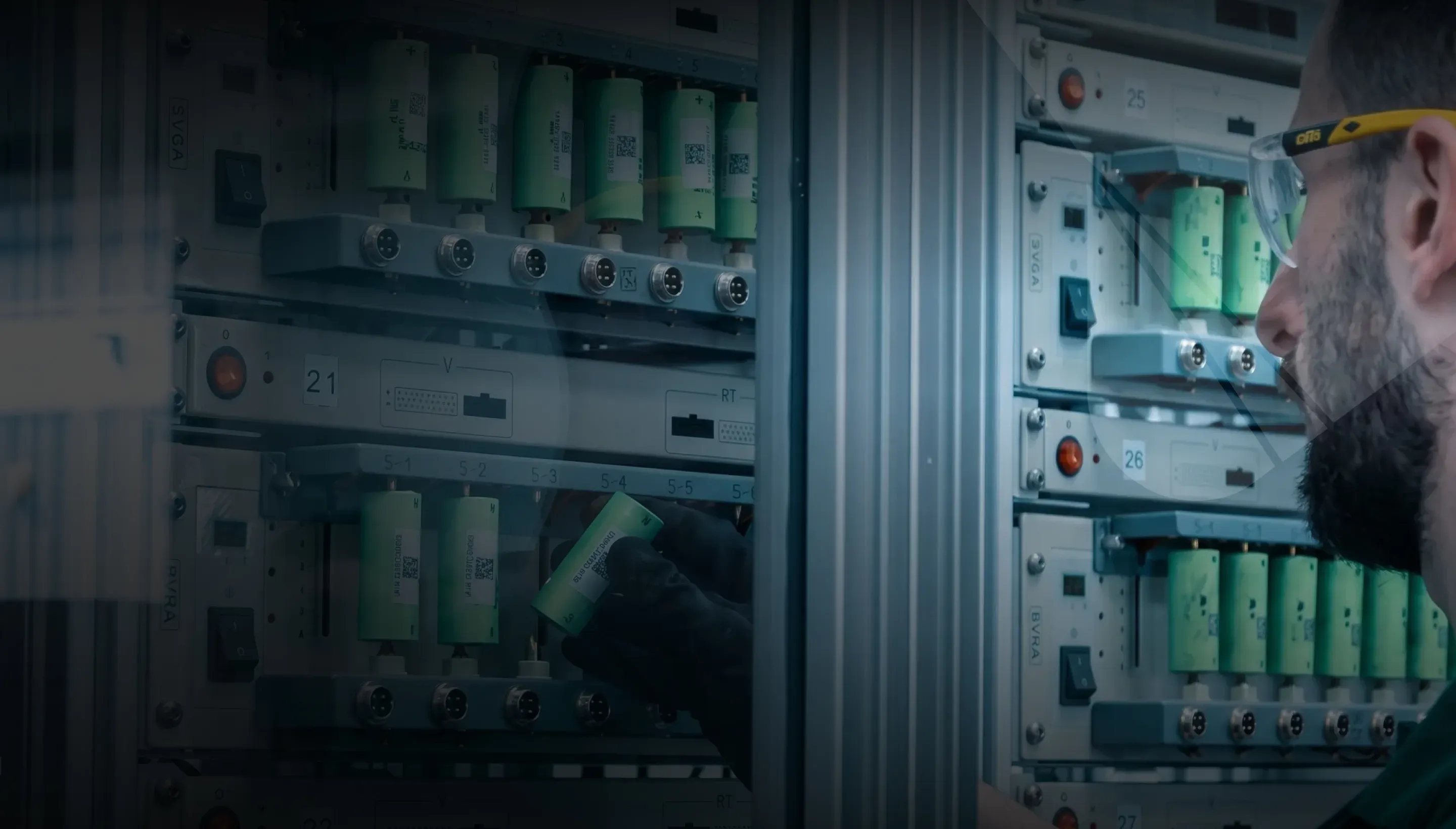 A technician in safety glasses working at a rack of green high-voltage battery modules